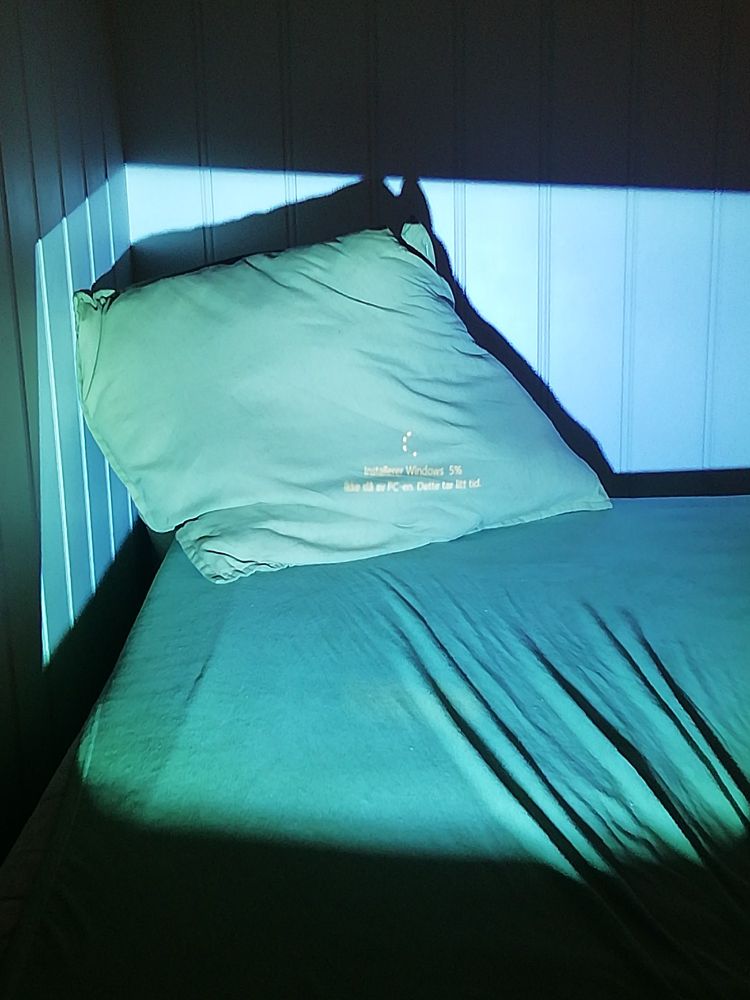 The corner of a bed with a pillow partly lid up by blue green light from a projector. On the pillow one can see a loading circle and read "Installerer Windows. 5% Ikke slå av pcen. Dette tar litt tid." The sheets are wrinkled in the bottom right corner creating lading lines from the text on the pillow.