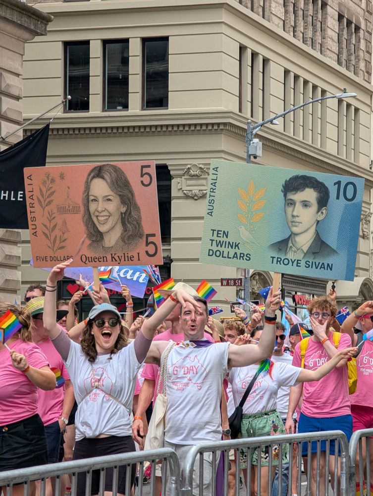 the Australia contingent of NYC pride holding up two signs meant to look like Australian currency: five dollars with Kylie minogue's face, and ten twink dollars with troye sivan