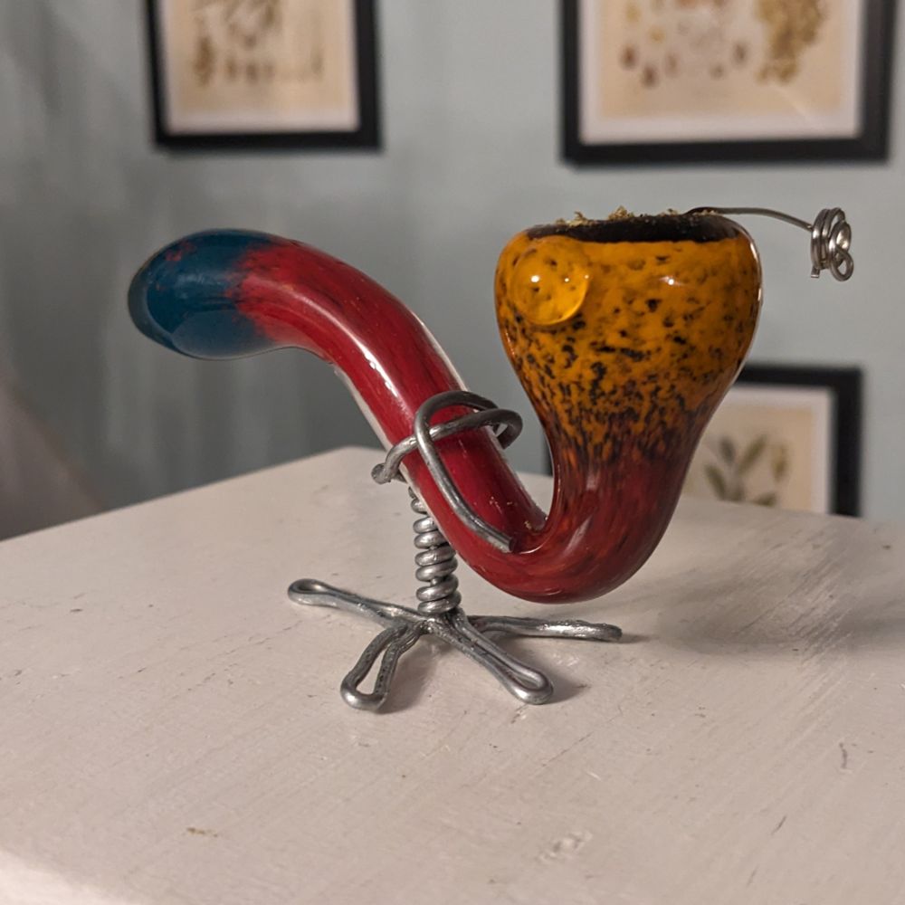 My new pipe, a cheap clearance sale red eye glass pipe with a home made stainless steel smoking screw and a galvanized steel wire art foot in a shape resembling a bird's foot with a center wrapped leg and crude wrap around the pipe at the balance point.