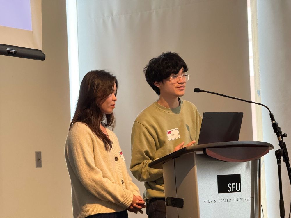 Jeffrey Wong and Arwen Fong presenting at the Northwest Robotics Symposium at Simon Fraser University. A laptop is open in front of them with an SFU sign on the lectern.