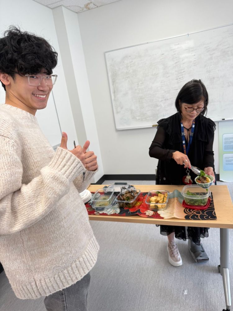 Jeffrey Wong smiles and gives a double thumbs-up beside Dr. Lillian Hung, who is serving food from a spread of Asian dishes including braised meat in curry, fried tofu, and leafy greens.