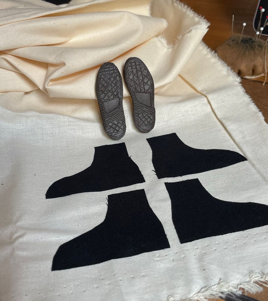 Na foto estão os moldes de tecido para a confecção dos tênis em miniatura em preto, mais atrás estão os solados feitos em cinza escuro e no fundo das peças tem um pedaço de algodão cru 