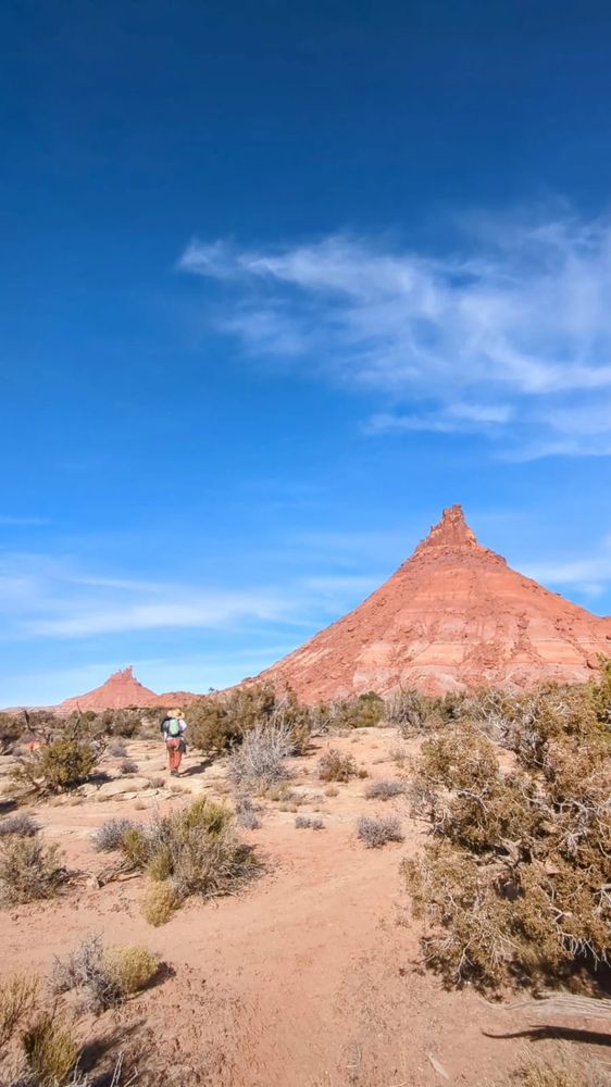 A vertical landscape showing two desert towers of red rock. A woman is walking towards the towers in the foreground with a straw hat and mint green backpack.