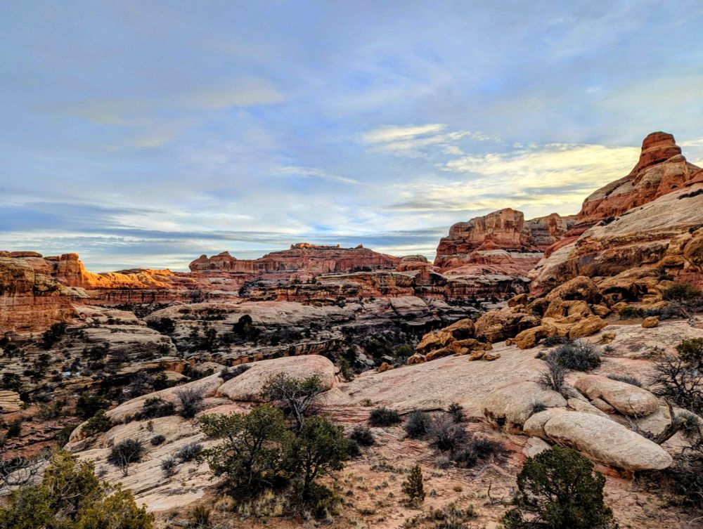 A landscape of the backcountry desert in Canyonlands National Park. The sky is blue with a few lingering clouds, and the rocks are straited with white and red layers. 