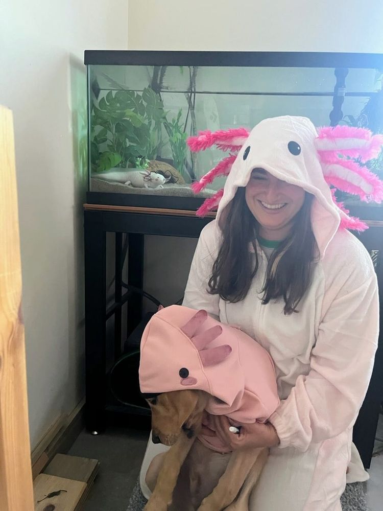 A woman and a labrador puppy, both in axolotl costumes, kneel in front of an aquarium. Inside is an axolotl, and dog ears have been drawn on the glass. The woman is smiling, the dog looks demoralized, and the axolotl betrays no emotion.