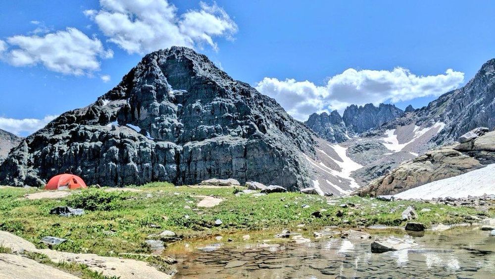 An orange tent pitches beside a lake, with a massive, cone-shaped peak in the backgroun.