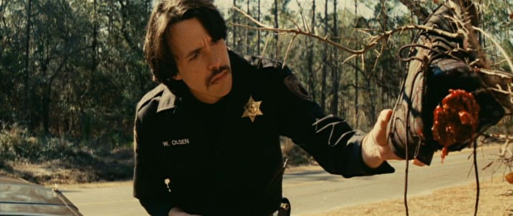Screenshot of Cabin Fever 2: Spring Fever from 2009: Deputy Winston, now he has no sunglasses and he gas a different haircut, touches a shoe caught in a tree that has Paul's dismembered foot still inside. In the background, there's the hood of his police car, the road, and a forest of longleaf trees.