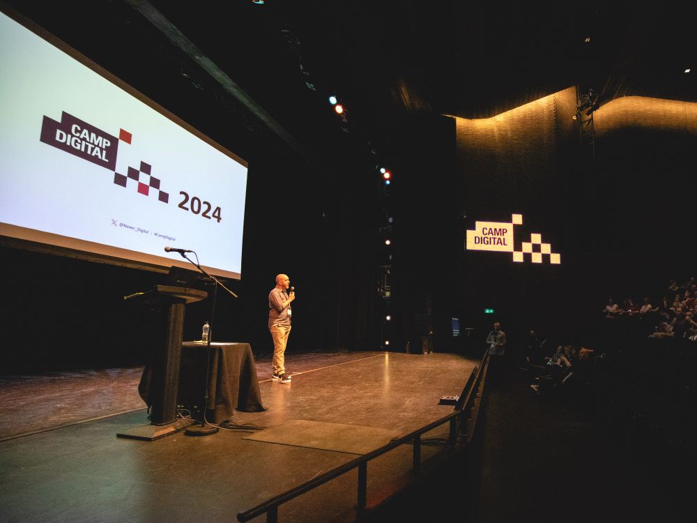 Camp Digital host Shaun Gomm onstage at the RNCM Manchest. The Camp Digital logo is seen on the slide behind Shaun, and projected onto the wall. 
