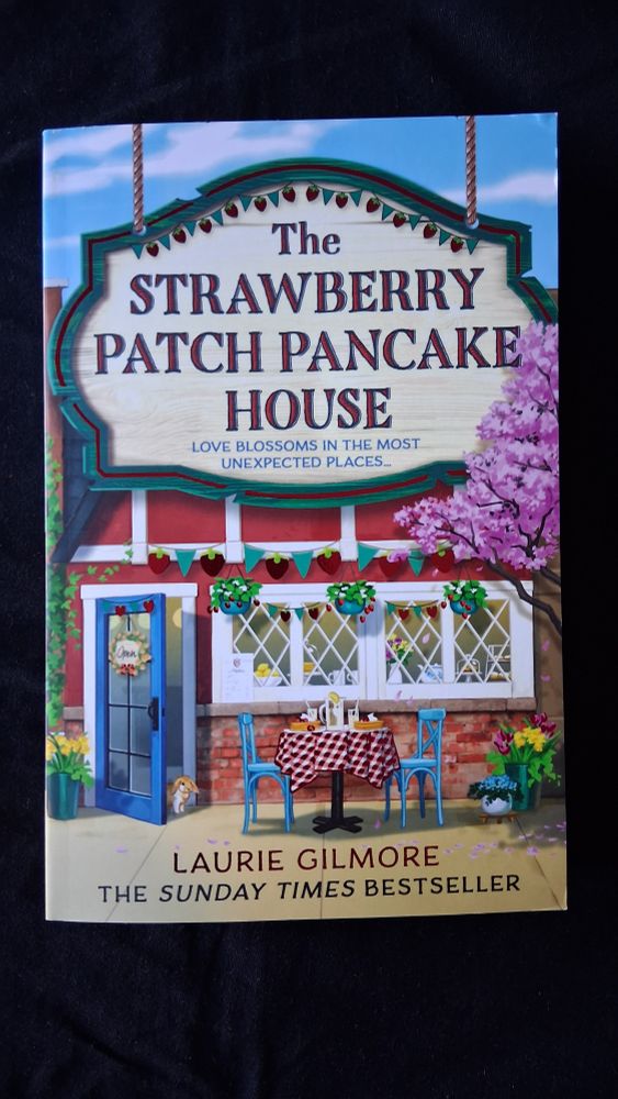 Book cover for The Strawberry Patch Pancake House by Laurie Gilmore, showing a shop front with a table and chairs set up outside for diners.