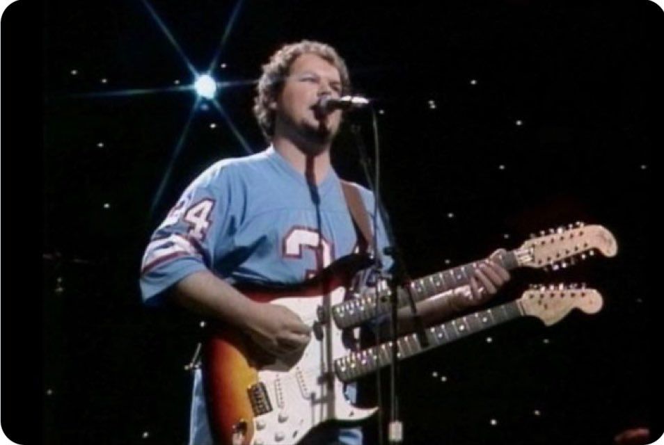 Christopher Cross wearing a light blue Earl Campbell Houston Oilers jersey while playing a double guitar. 