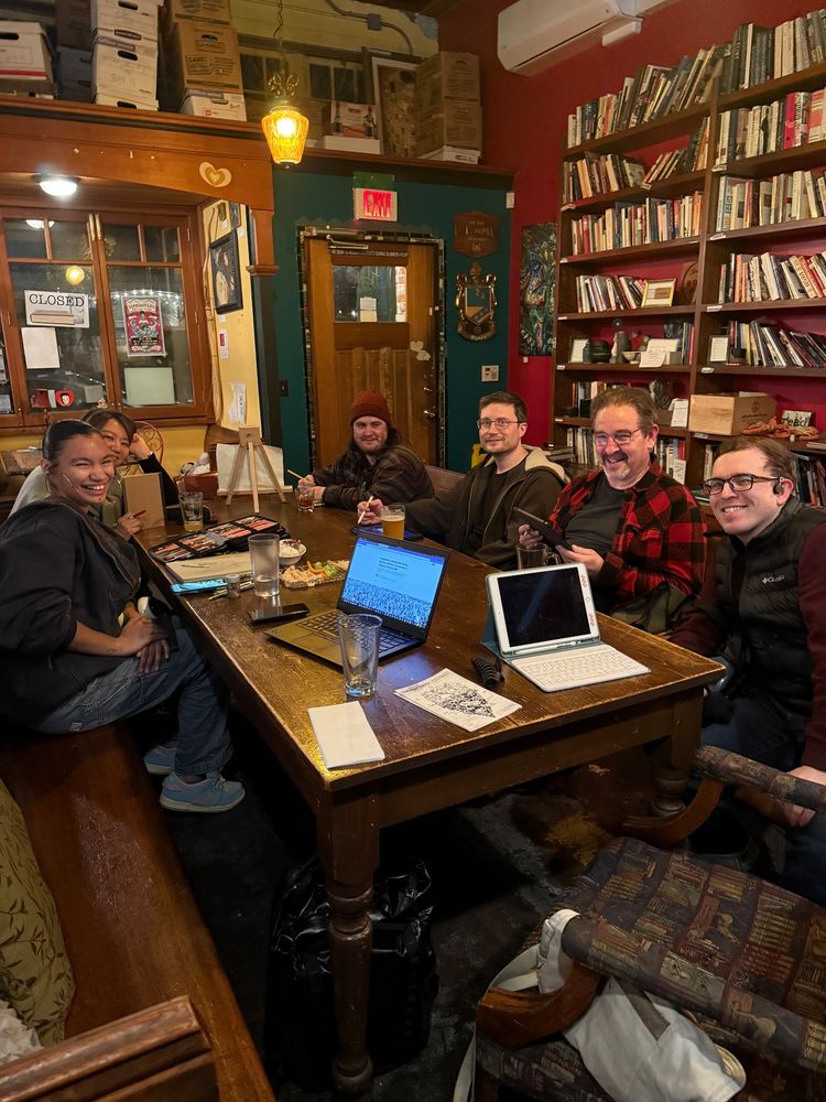 Men and women sitting around a table smiling with laptops, tablets and physical art supplies. A large bookshelf can be seen in the background of a local book pub.