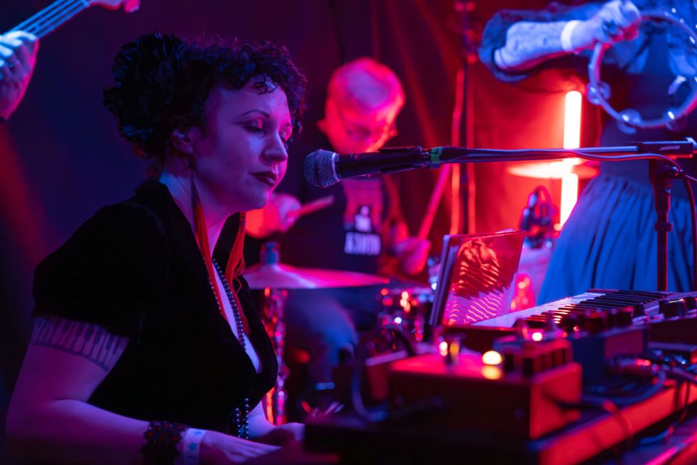 Unwoman, who has pale skin, black curly hair, and goth style, is lit in red and blue, seated playing keyboard on stage at the upstairs of DNA Lounge. Sergey the drummer and Andrei the bassist and Rebecca the lead singer are partially visible. 