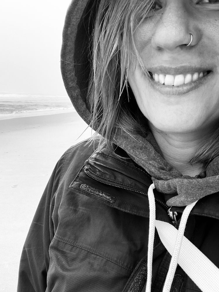 Eine schwarz-weiß Nahaufnahme von mir. Ich trage einen Hoodie und darüber eine Jacke. Die Kapuze des Pullis habe ich auf dem Kopf und lächle. Ein paar Haarsträhnen fallen mir ins Gesicht. Hinter mir sind verschwommen Meer und Strand im Nebel zu erkennen. 