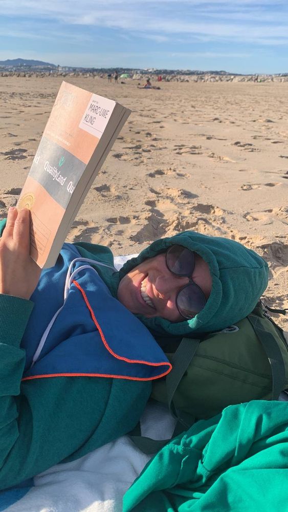 Ich liege mit hoodie am Strand und lese. Außerdem bin ich mit einem Handtuch zugedeckt, das ich mir bis unters Kinn gezogen habe. Im Hintergrund Strand, blauer Himmel und, nun ja, in der Ferne Menschen in Badesachen, denen es scheinbar nicht kalt ist. 