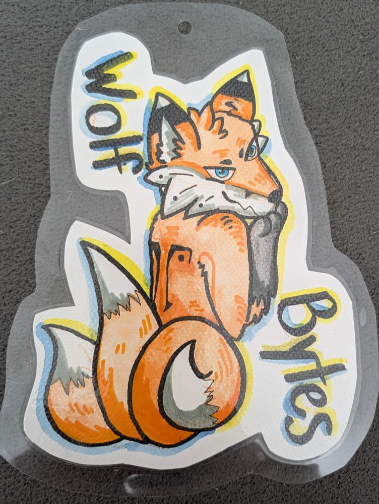A drawn laminated badge of a three-tailed kitsune looking over his shoulder mischievously, with the name "Wolf Bytes" added.