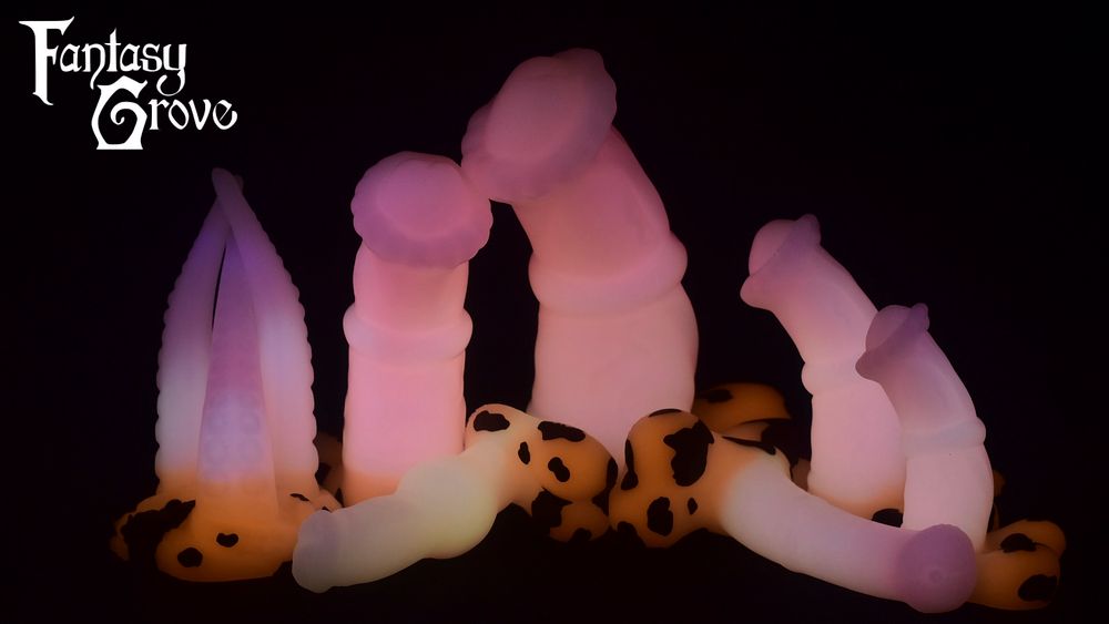 A lineup of dildos showing off their glow in the dark. Glow is a darker pink to lighter pink fade on the shaft, and orange glow on the base with dark brown cow spots. From left to right there's a three shafted tentacle model, a large chunky equine, a XXL chunky equine, and a medium and small chunky equine. In the front is a canine model and a longer thinner equine model.