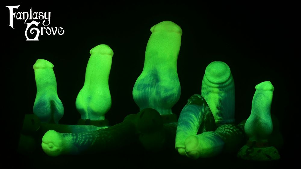 A lineup of dildos in the dark with green to blue ribbony glowing shafts. The Amaroks have hand painted collars and chains and many models have hand painted sprinkles, some of which can be seen faintly backlit by the glow of the toy. From left to right there's a small, medium, and large knotted model (Amarok), a girthy humanoid, and a XS Amarok. In front laying down are a realistic humanoid, a lengthy equine, a dragon packed, and a knotted realistic werewolf.