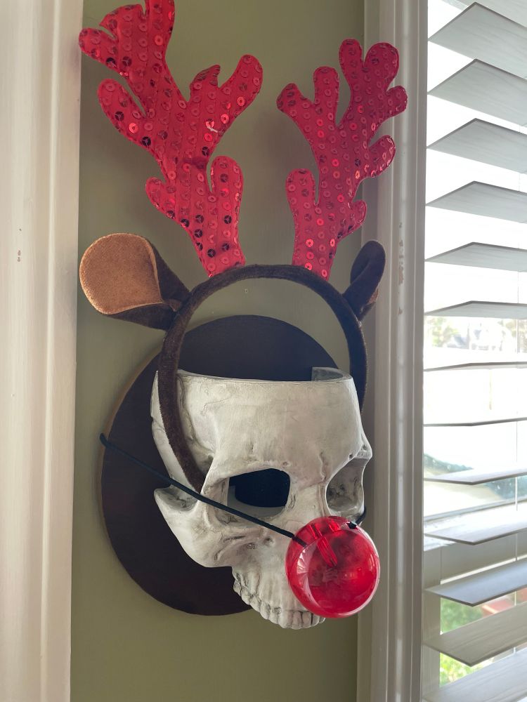 A mounted human skull hangs on a wall with felt reindeer antlers and a light-up red nose.
