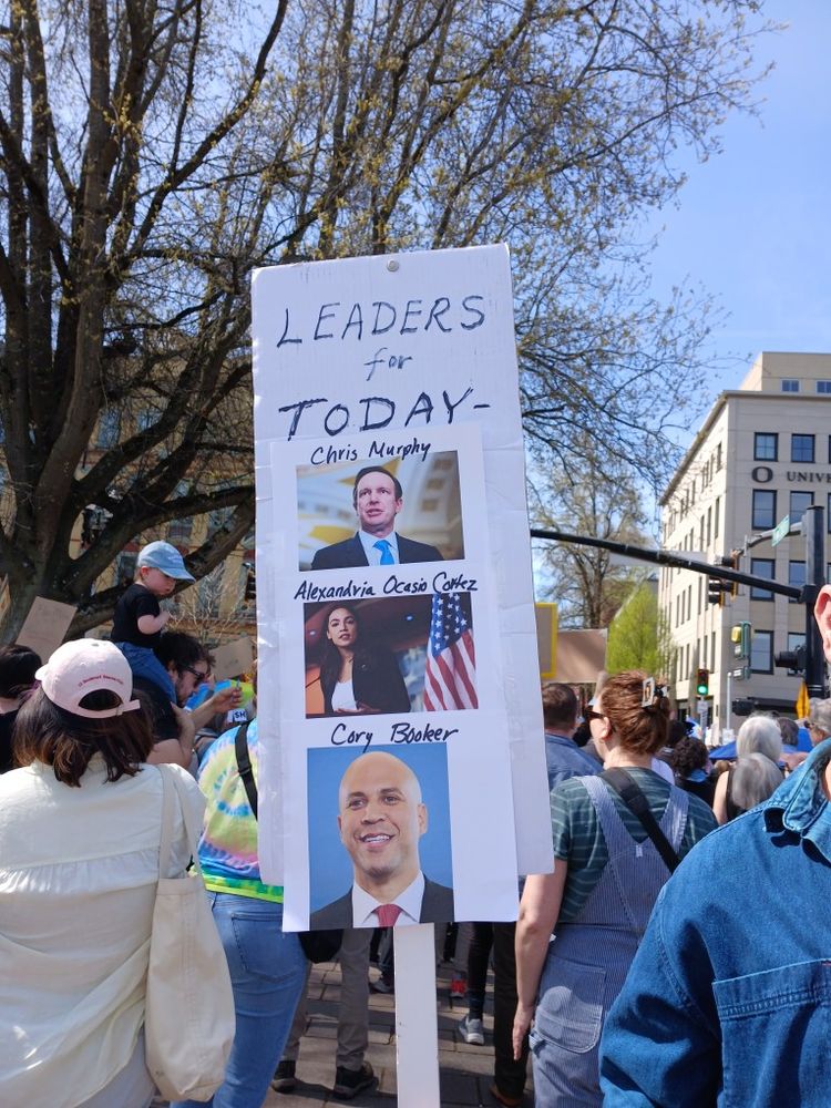 Amidst a crowd of protestors, one sign reads, "Leaders for Today - Chris Murphy, Alexandria Ocasio Cortez, Cory Booker," with corresponding photographs. 