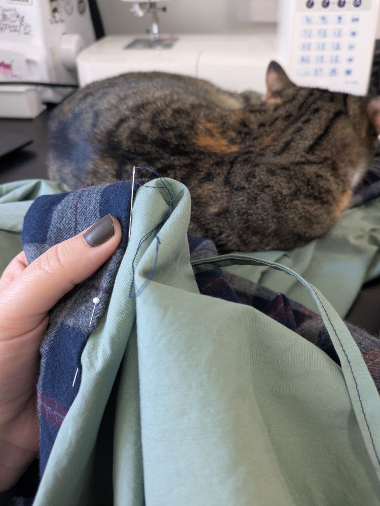 A hand holding up fabric being hand stitched together. One later is a navy plaid and one is a sage green. The background is a tabby cat having a little nap on the other end of the fabric. As one does. 