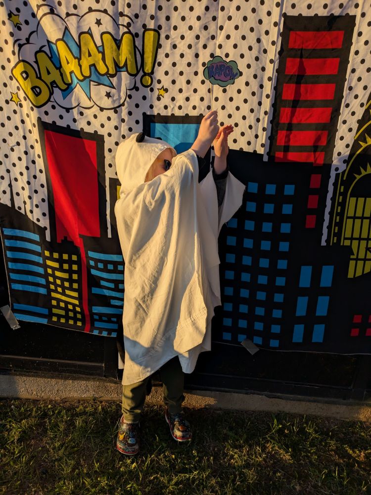 A small child in a white ghost cape and a black cat tail and ears stands in front of a pop art style backdrop with their arms up