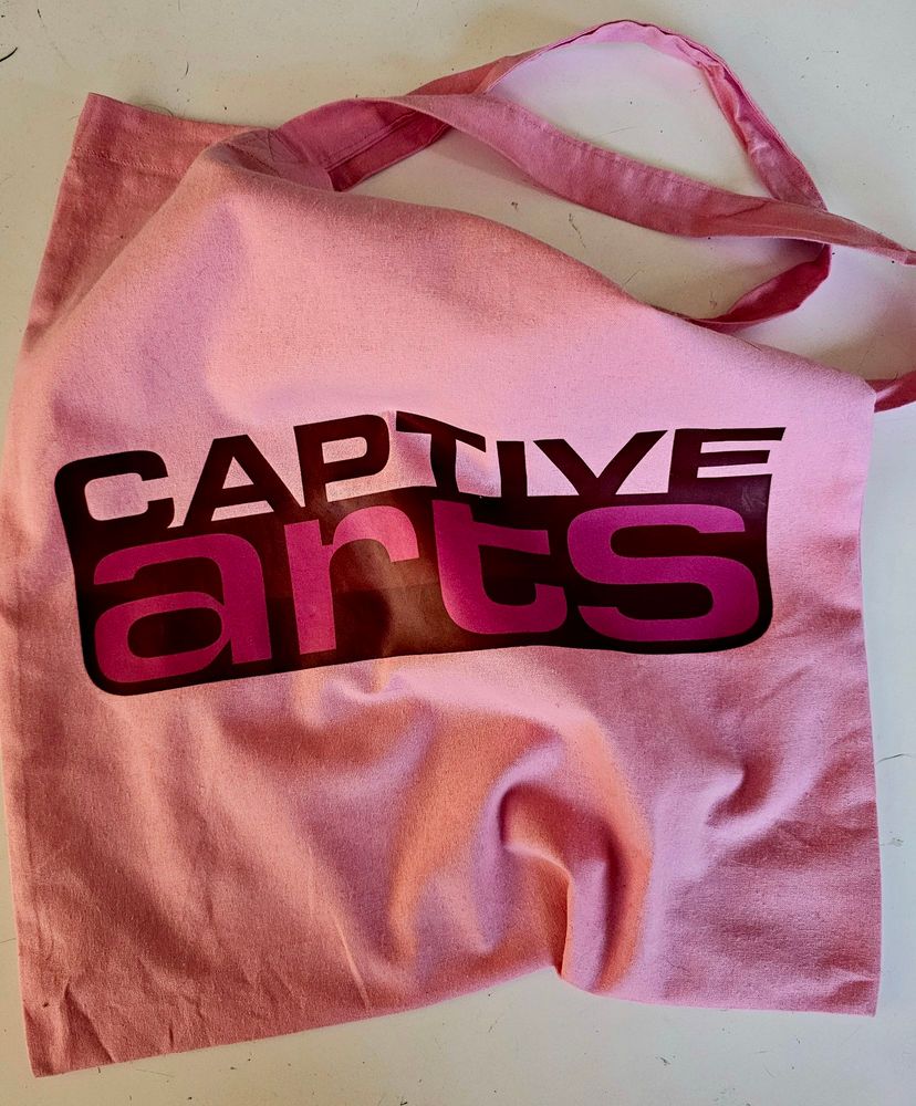 An empty pink tote bag, slightly crumpled, on a white surface. With the words CAPTIVE Arts printed on the front. 