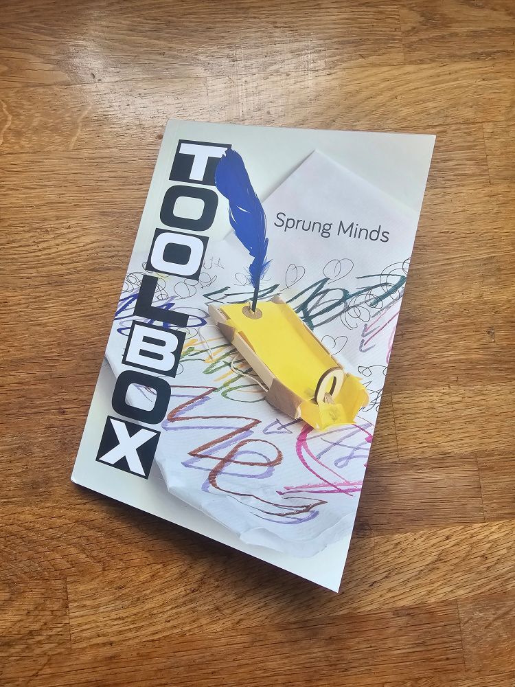 A book on a wooden surface. The cover title in black is 'Toolbox: sprung minds'. The ground image is a rudimentary yellow boat with a blue feather sail on a sea of doodles 