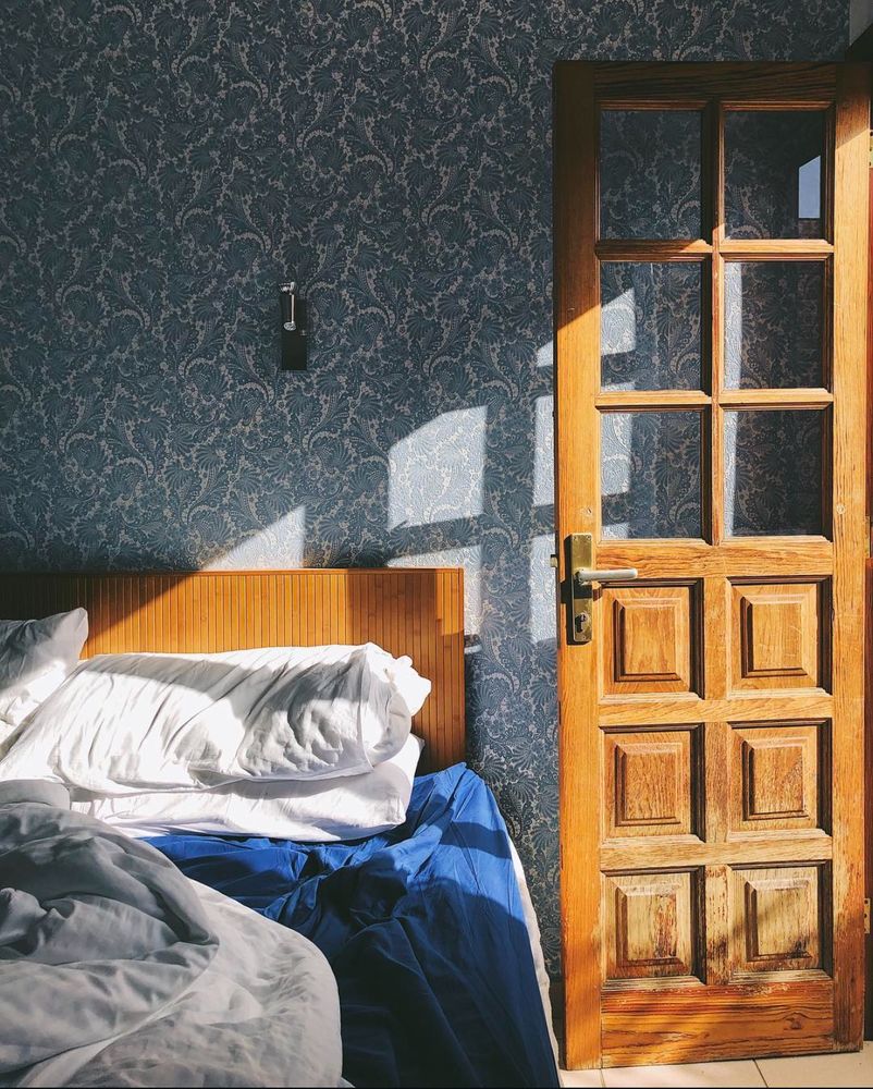 A sunlit bedroom featuring a wooden bed with white pillows and a blue bedsheet, adjacent to a wooden door with glass panels. The wall has a patterned blue wallpaper, and natural light streams through the door, creating soft shadows on the bed and wall.