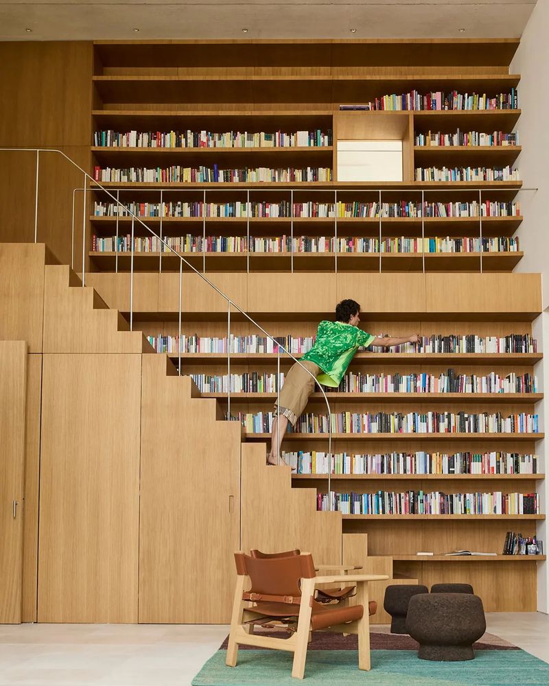 una estantería de dos plantas de altura atravesada por una escalera. En medio, un hombre intenta acceder a un libro en un extremo inaccesible del estante superior
