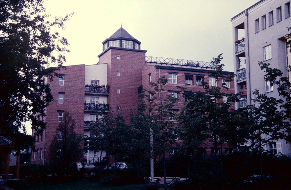 Vista trasera del edificio. El bloque prismático se rompe desde este alzado donde destaca un cuerpo en forma de torreón, más alto que el resto del edificio y que alberga la comunicación vertical de las viviendas. El torreón está rematado por una cubierta metálica poligonal y un barandilla metálica con listones cruzados en x. Todo el edificio está revestido de ladrillo, excepto un cuerpo junto al torreón que alberga balcones
