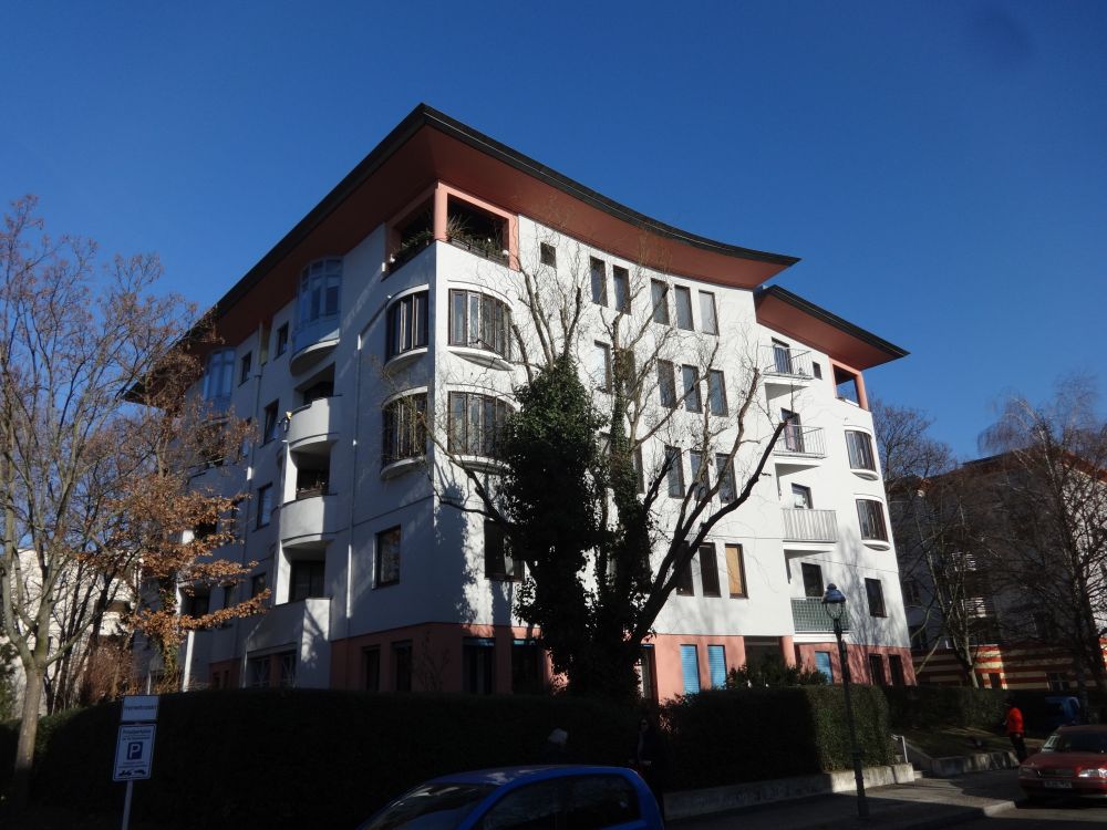 Imagen de una villa urbana de apartamentos en Berlín. El edificio de volumen cúbico y estilo posmoderno tiene una gran cornisa plana, balcones en esquina, ventanas mirador circulares atravesando una esquina y planos de fachada rotos para introducir un balcon oblicuo.