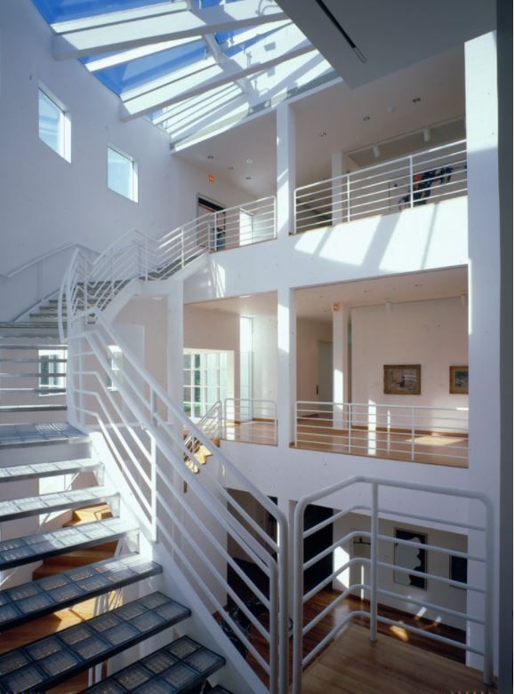 Vista del Interior del museo. A la izquierda, una escalera de peldaños sin contrahuella y piso de bloques de vidrio, asciende bordeando un muro semicircular rematado por una alienación de ventanas cuadradas y cubierto por una claraboya con un estructura de grandes perfiles en abanico pintados de blanco. La barandilla de la escalera es de tubos metálicos pintados también en blanco. el fondo se ve un gran espacio de tres alturas, abierto a las tres plantas de galerías donde se exponen obras.