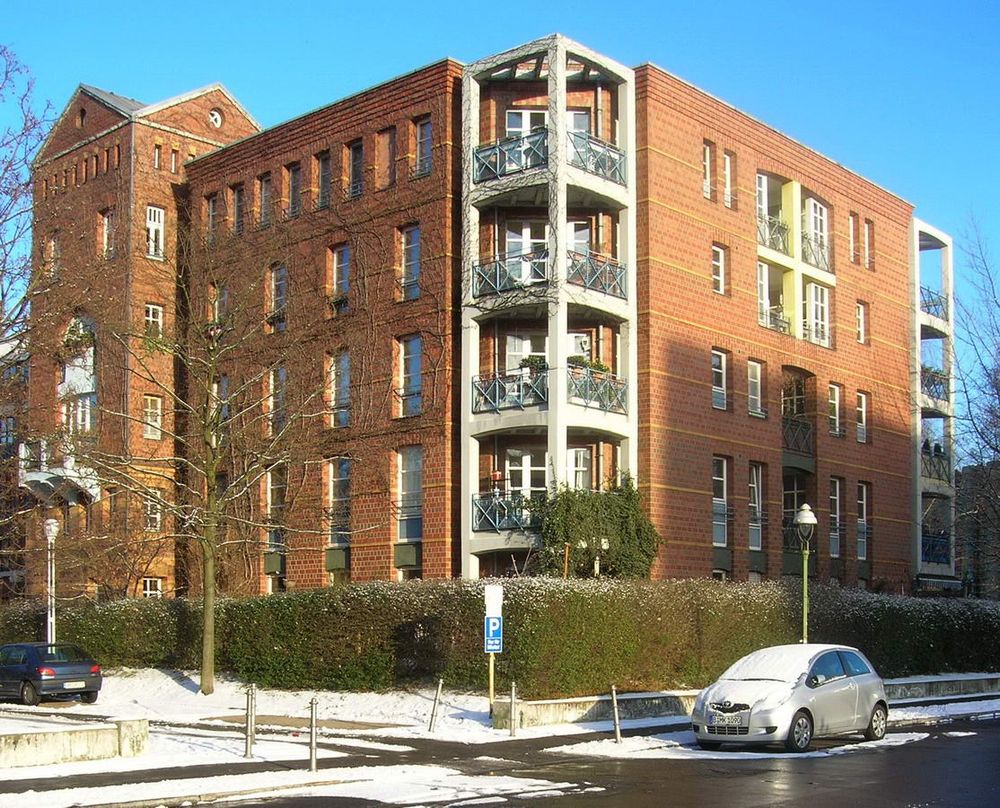 Esquina del edificio de viviendas. tiene fachada de ladrillo, un torreón con cubierta inclinada y las esquinas tiene balcones triangulares construidos sobre una estructura de hormigón claro que contrasta con el edificio por su material y que además parece separada de él.