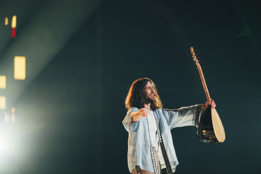 Louis Desseigne, a man with long brown hair and a beard, holds a bağlama aloft
