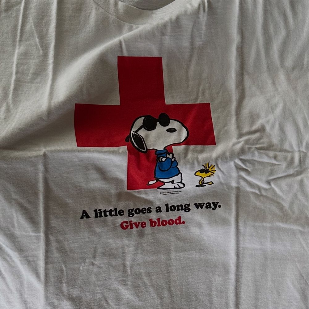 Detail of a white tshirt with a big Red Cross on it. Snoopy and Woodstock stand by the cross. The text below reads “A little goes a long way. Give blood. 
