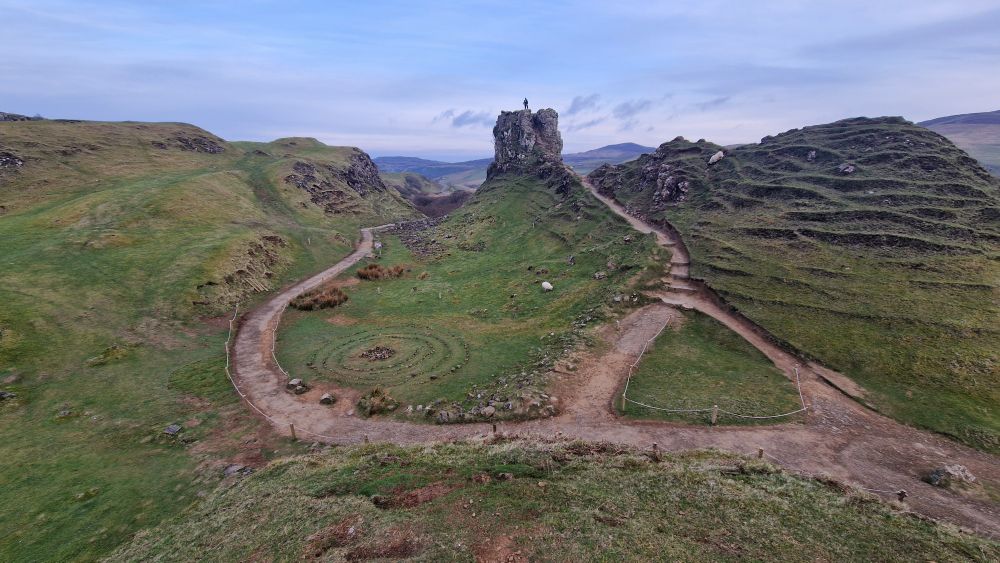 Photo of the fairy glen on Skye, with pathways leading up to a basalt tower outcrop and a person standing on top