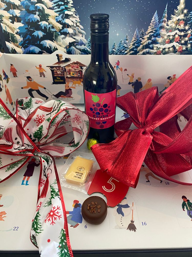 Small glass bottle of red wine with fuchsia label posed between two large ribbon bows with a plastic wrapped yellow cheese and round chocolate bonbon
