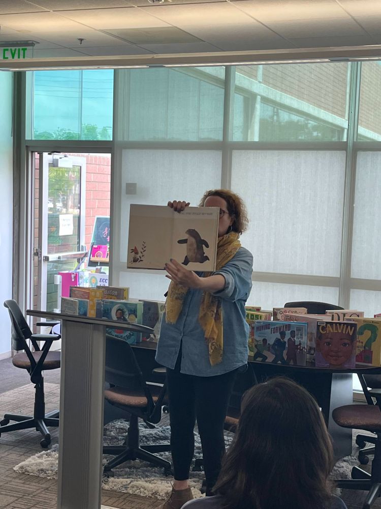 A woman shows the interior of a picture book to an audience, with a display of other picture books behind her