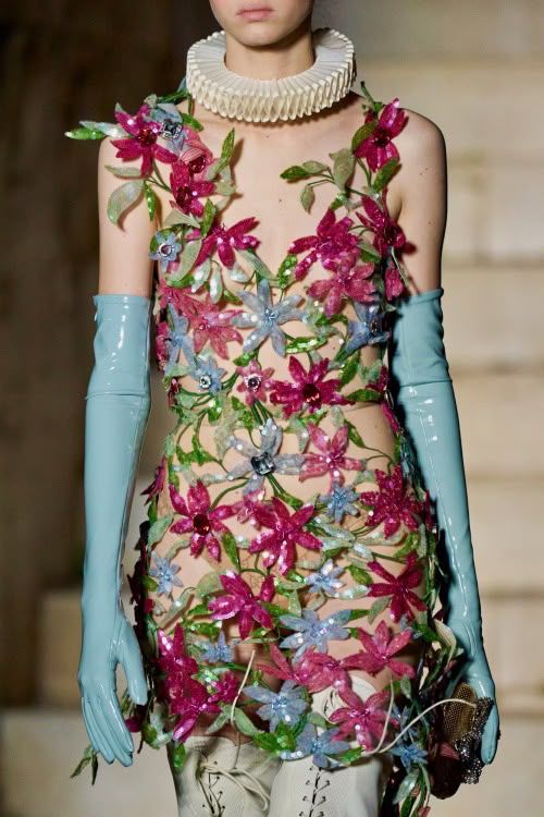 A model wearing a bold floral dress made entirely of three-dimensional pink, purple, and blue flower appliqués connected by delicate green vines. The sheer ensemble is paired with pale blue latex gloves and a structured ruffled collar, blending romance with futuristic elegance. Gucci 2023