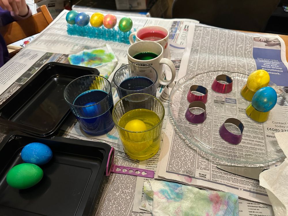 Eggs in various stages of being decorated: some soaking in dye cups, some drying, some waiting for gold paint to be applied over the dye, and some sitting in shiny display rings 