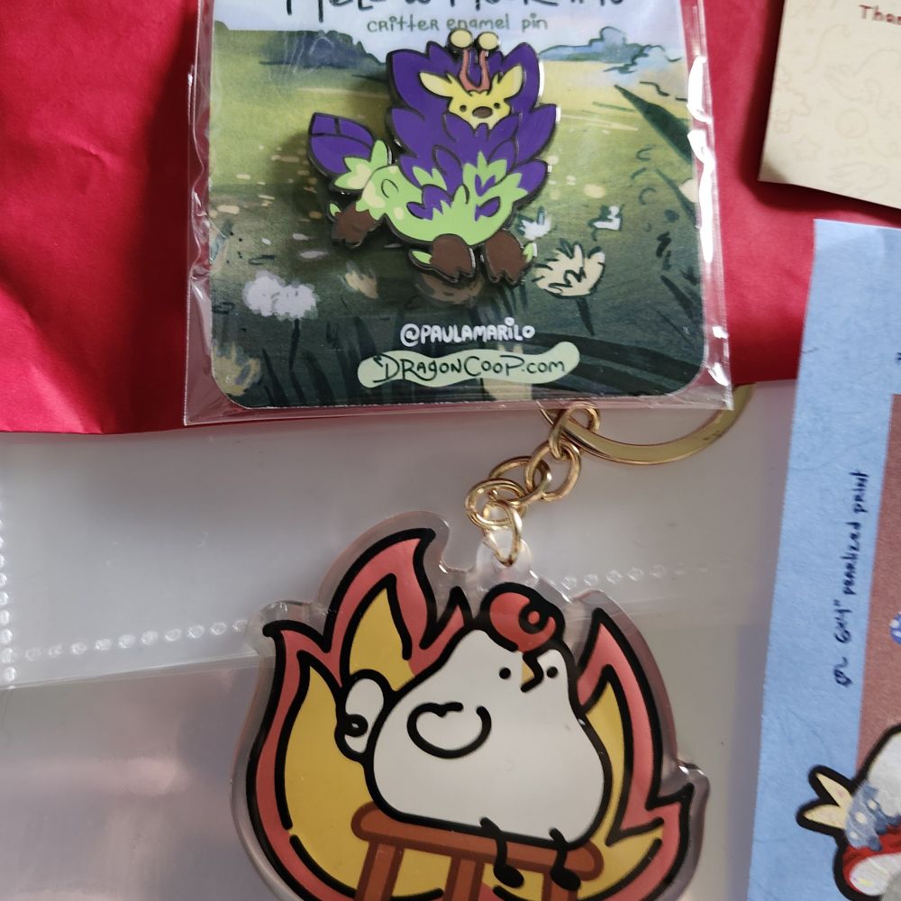 A pin of a green and purple mossy dragon and a charm of a chicken sitting on a stool with a fiery background.