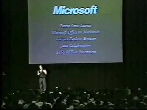 Steve Jobs standing on stage at Macworld 1997 announcing Apple's partnership with Microsoft. On the screen behind Jobs is the Microsoft logo, along with the 5 aspects of the partnership. They read "Patent Cross License, Microsoft Office on Macintosh, Internet Explorer Browser, Java Collaboration and $150 Million Investment."