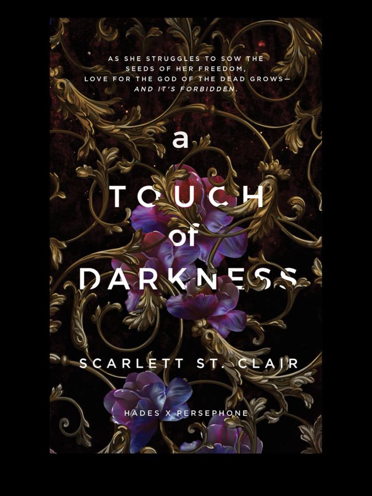 The book cover for *A Touch of Darkness* by Scarlett St. Clair features an intricate design with gold ornamental vines weaving across a dark background. Among the vines, vibrant purple and blue flowers bloom, adding a touch of color and elegance. The title is displayed in bold white capital letters, partially entwined with the floral elements. Above the title, a tagline reads: "As she struggles to sow the seeds of her freedom, love for the God of the Dead grows—and it's forbidden." The author's name appears below the title in white text, along with the note "Hades x Persephone," indicating a retelling of the mythological love story.