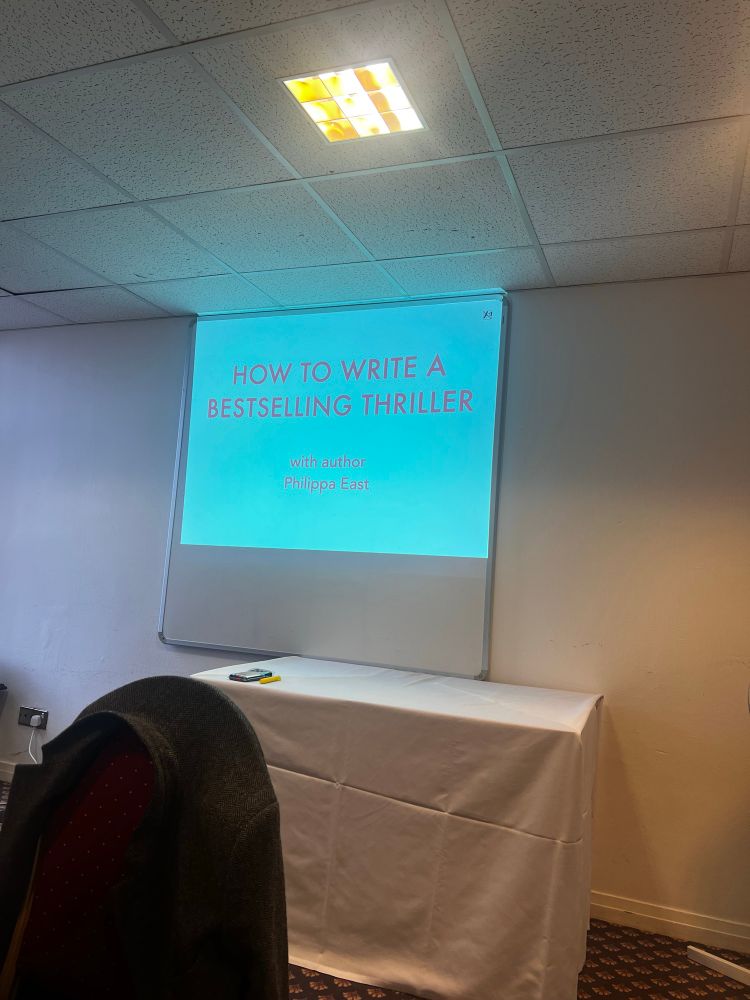A presentation slide projected onto a white screen in a conference room. The slide has a blue background with red and white text that reads, "HOW TO WRITE A BESTSELLING THRILLER with author Philippa East." Below the screen, a table covered with a white cloth holds a remote control and a yellow highlighter. In the foreground, a red upholstered chair with a dark coat draped over it is visible. The room has a white ceiling with fluorescent lighting and beige walls.