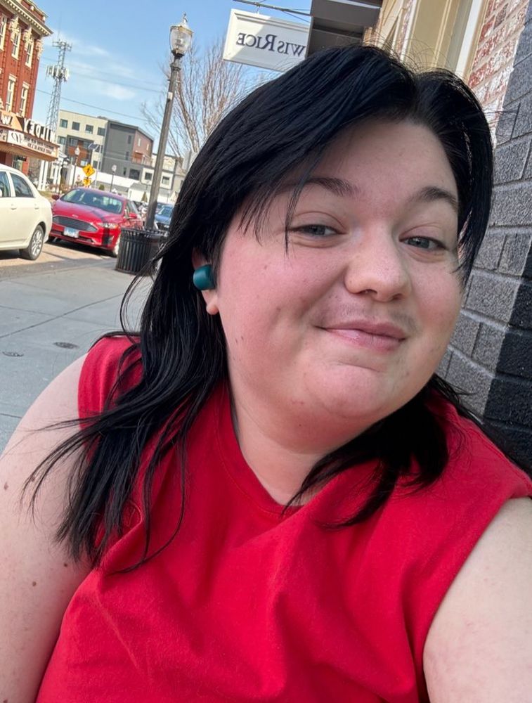 another selfie, 6 months on testosterone taken today in 2025. my hair is past shoulder length in an overgrown mullet, dyed black, i’ve got some semblance of a mustache, i’m wearing a red bro tank, and have gained some weight since the last photo. my face has become slightly more masculine as well