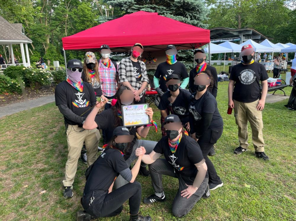 12 JBGC members posing in a park in front of some pop-up canopies, holding a certificate of appreciation from Woonsocket Pride