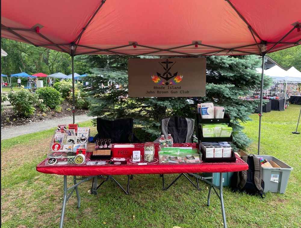 A table in a park under a red pop-up canopy, stocked with fundraising items and info and harm reduction supplies. A banner hanging from the back identifies it as belonging to the Rhode Island John Brown Gun Club