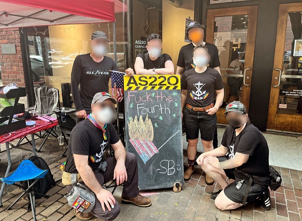 Six community defenders pose around a chalkboard from AS220. The chalkboard says "Fuck the Fourth" above a drawing of a burning US flag. It's signed "SB." a torn-up US flag hangs from the chalkboard.