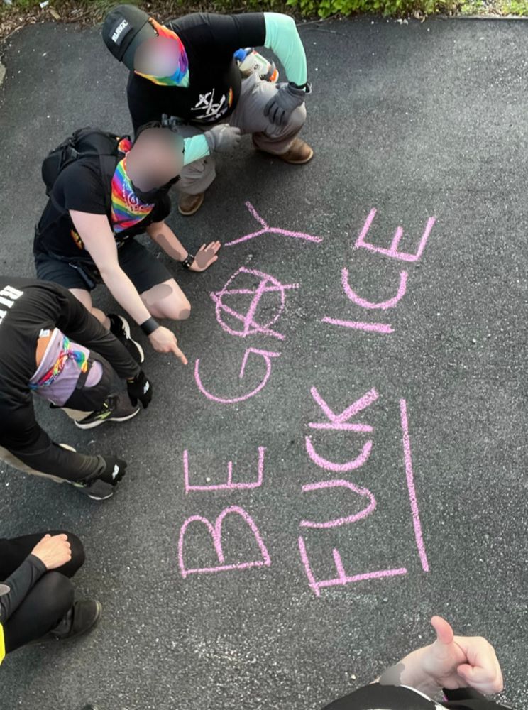 5 RIJBGC community defenders, most partly out of frame, crouch around a chalk slogan on blacktop reading:
BE GAY
FUCK ICE

The A in GAY is the anarchy A and FUCK is underlined