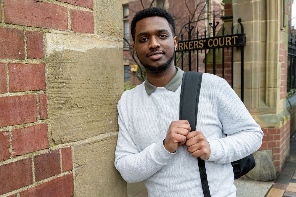 Dawit leans against the wall, wearing a rucksack over his shoulder. He is stood in front of Clothworkers' Court gate at the University of Leeds